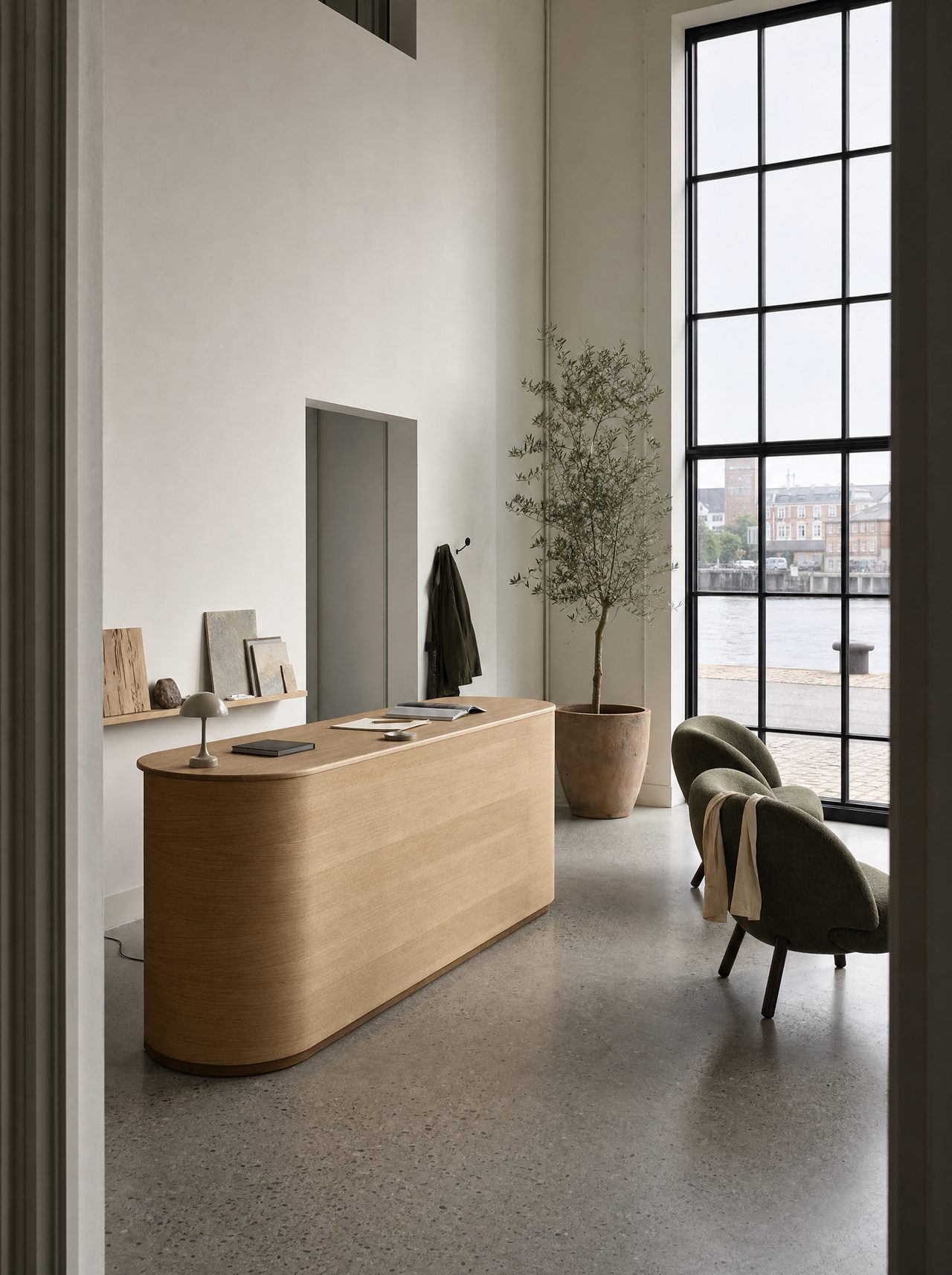 The front room of a small modern business on the Copenhagen harbour on a grey morning: a rounded oak counter, two olive wool chairs, an olive tree and a tall steel window onto the quay.