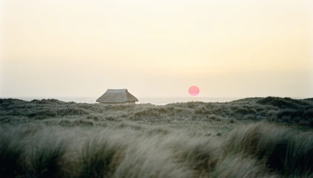 A thatched summer house in the dunes at sunset, with a low red sun on the horizon.