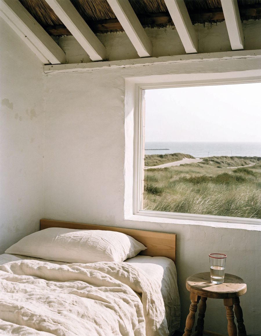 A summer house bedroom under white beams, linen on the bed and a window onto dune grass and the sea.
