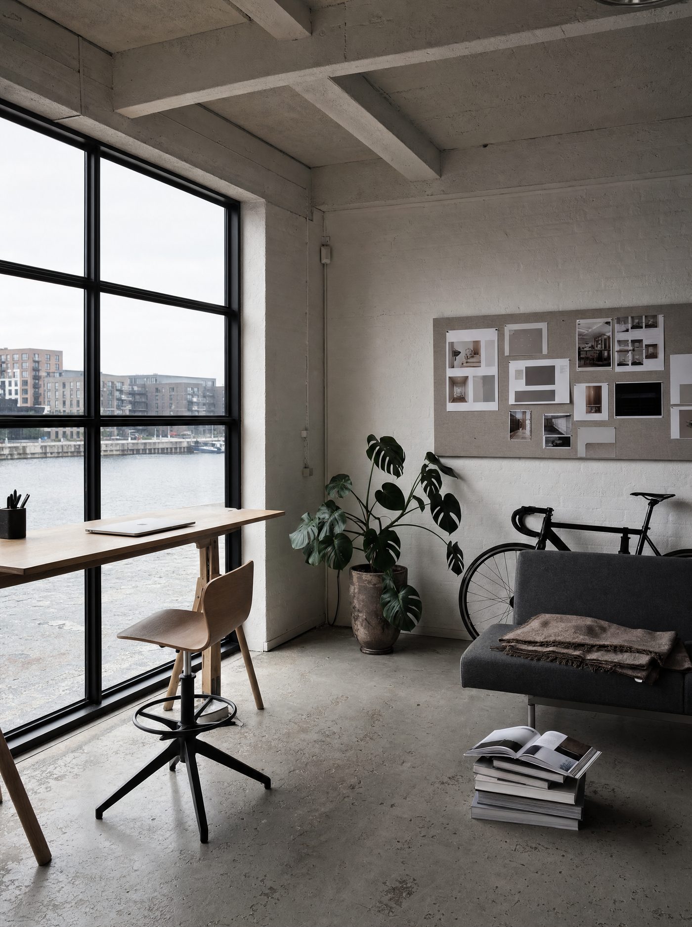 The other end of the same studio: a standing desk by the window over the harbour, a pinboard of printed website layouts, a grey sofa, a stack of design books and the bike.