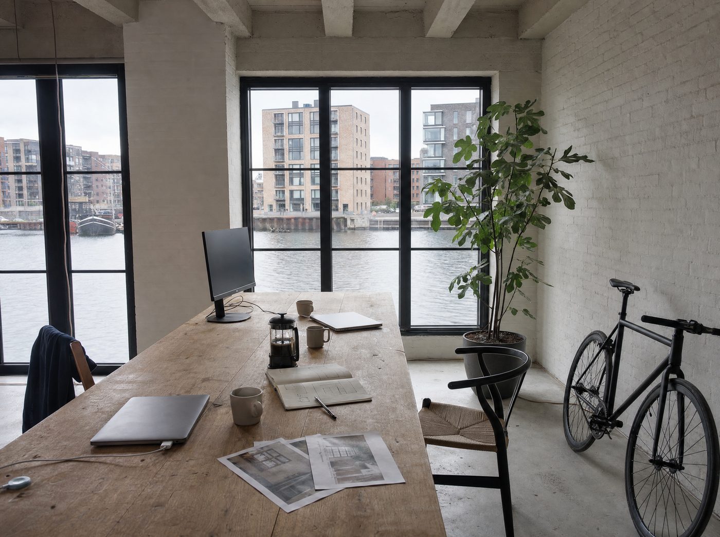 Our kind of studio, on the top floor of a harbour warehouse in Nordhavn: a long worn oak table with closed laptops, sketches and a coffee press, a fig tree and a bike by the steel windows.