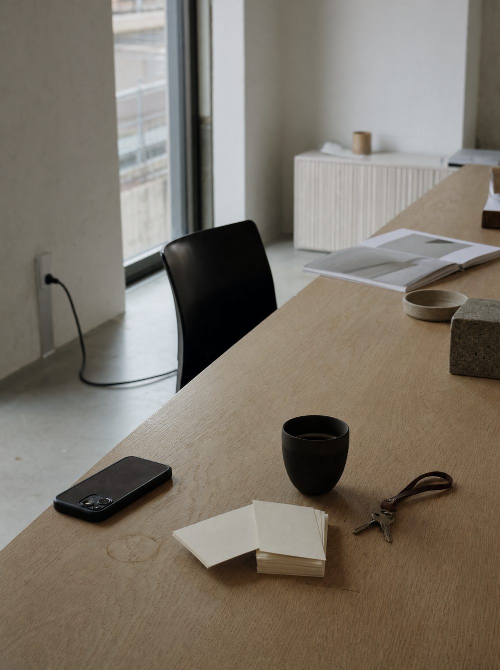 An oak counter on an overcast morning: a phone lying face down, a loose stack of blank cards, a black coffee and a set of keys.