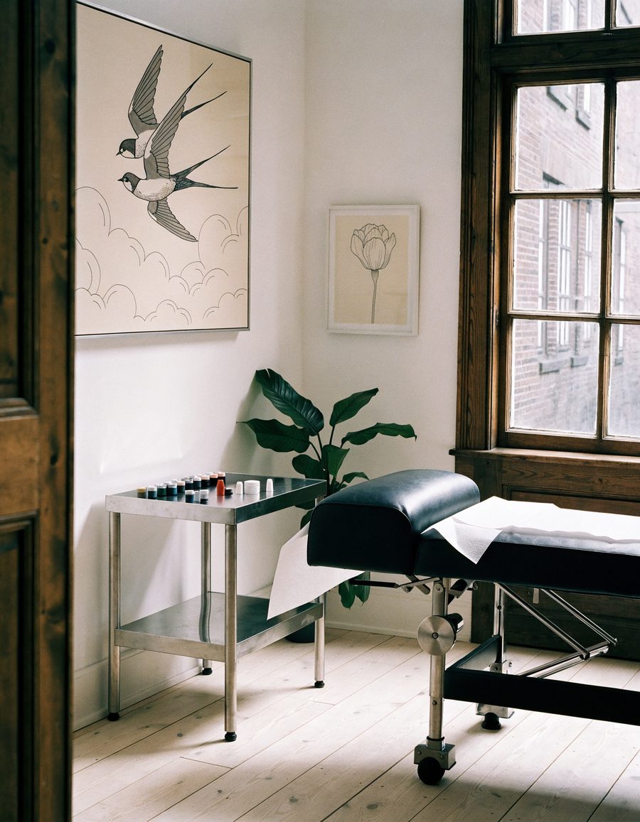 A calm tattoo studio: a black leather bench by a tall window, a steel trolley of inks, swallow drawings on the wall.