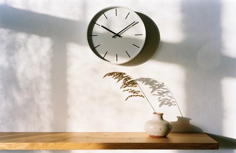 A plain white wall clock above an oak shelf, in afternoon sun.